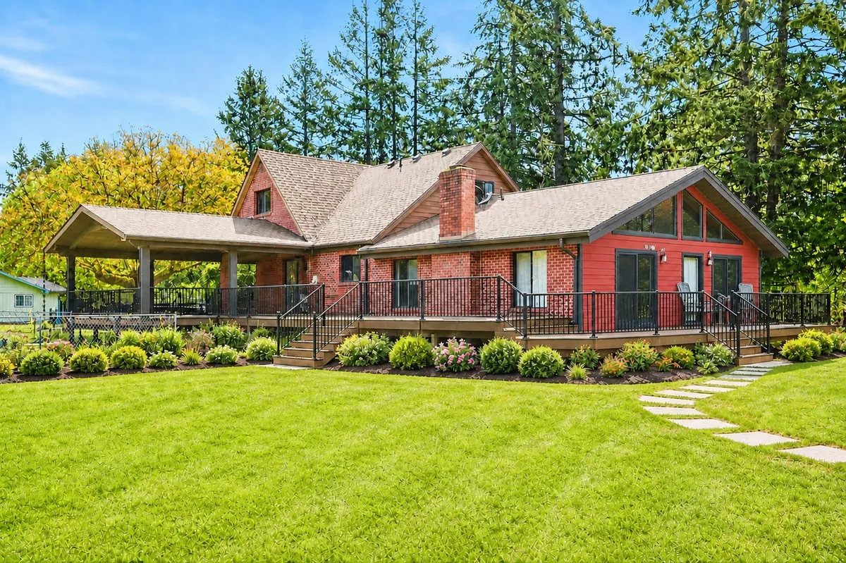 A red brick ranch house with a new wraparound deck and covered patio, a wide green lawn and fir trees behind